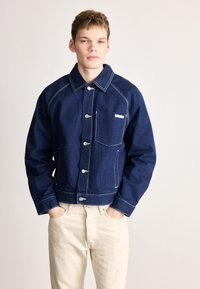 Dongerijakke i mørk blå med hvite sømmer, med knappelukking, to frontlommer og en strukturert krage. Kombinert med lysebeige bukser.