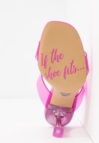 Sandale à talon haut transparente rose montrant la semelle avec le texte "If the shoe fits" en cursive, positionnée contre un fond blanc en coin.