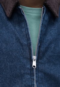 Åpen blå denimjakke med brun kordfløyelskrage over en grønn ribbestrikket skjorte på brun hud.