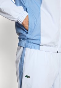 Ανοιχτό μπλε φόρμα με ελαστικές μανσέτες και μέση, κατασκευασμένη από υφή υφάσματος. Διακριτικό πράσινο λογότυπο κροκόδειλου στην αριστερή πλευρά του παντελονιού.