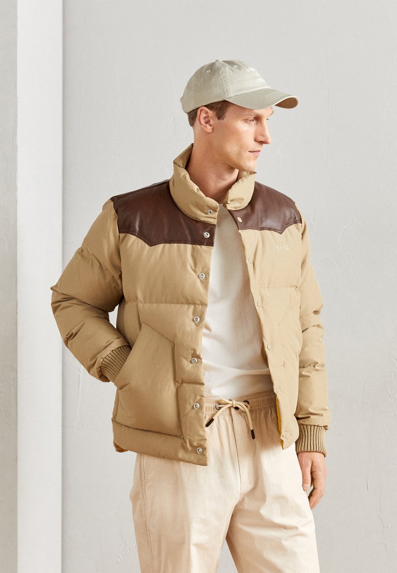 Giacca puffer beige con yoke in pelle marrone, bottoni a pressione e polsini a costine. Indossata sopra una maglietta bianca e abbinata a pantaloni crema.