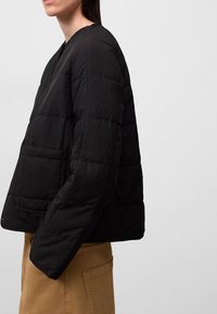 Giacca nera imbottita con scollatura rotonda, caratterizzata da un design trapuntato e tasche laterali, indossata sopra pantaloni beige.