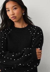 Sort sweater med rund hals, prydet med sølvdekorationsdetaljer spredt over skuldrene og ærmerne, strikket tekstur og lange ærmer.