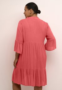 Mujer con el cabello oscuro recogido en un moño, vestida con un vestido suave color coral con mangas acampanadas y un dobladillo con volantes, de pie contra un fondo claro.