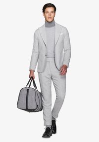 Grå ulddragt med matchende turtleneck, der har en subtil tekstur og slim fit. Manden bærer en duffelbag med fiskebensmønster.