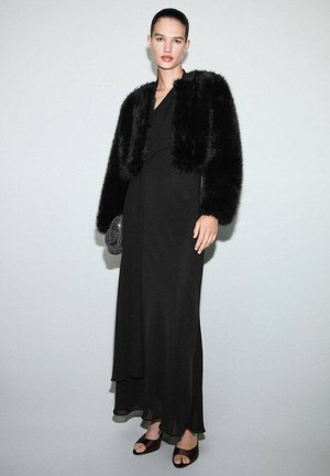 Vestido de noche negro con una silueta fluida, combinado con una chaqueta corta de peluche negra. Los accesorios destacados incluyen un pequeño bolso brillante y tacones abiertos.