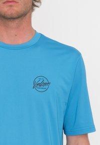 Camiseta de manga corta azul hecha de material suave con un logo circular negro que incluye el texto "Volcom" y la fecha de fundación en la parte frontal.