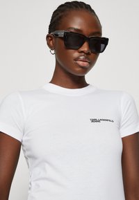 Biele priliehavé tričko s krátkymi rukávmi, na ľavej strane s čiernym logom "Karl Lagerfeld Jeans". Čierne slnečné okuliare.