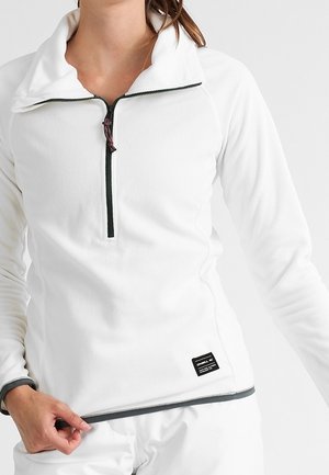 Nainen, joka käyttää valkoista pitkähihnaista fleecetakkia, jossa on musta puolivetoketju ja pieni musta etiketti helman lähellä, säätää takin alareunaa.