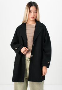 Young person with long, straight hair wearing a loose black buttoned coat, beige ruched top, and light green pants against white background.