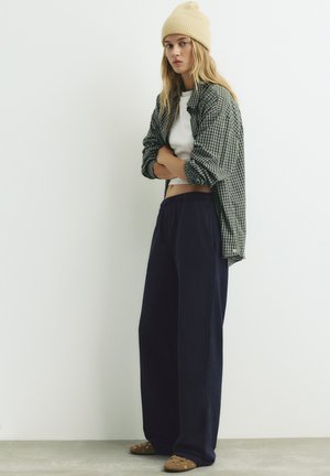 Ležerna odjeća koja uključuje košulju kariranog uzorka, bijeli crop top, tamnoplave široke hlače i smeđe cipele bez vezica sa dekorativnim zakovicama.