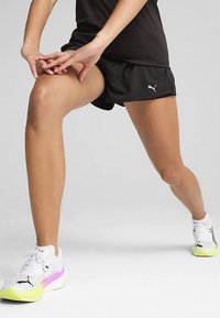 Juodi sportiniai šortai su PUMA logotipu, dėvimi su baltais sportbačiais, kurių vienintelis yra geltonos ir rožinės spalvų. Asmuo atlieka tempimo pratimą.