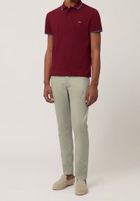 Bordeaux poloshirt met blauwe accenten, gecombineerd met lichtgroene broeken en beige instapschoenen, met een klein blauw logo op het shirt.