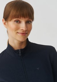 Modra sportska jakna na zatvaranje s visokim ovratnikom, s suptilnim logotipom i glatkom teksturom, dizajnirana za udobnost i performanse.