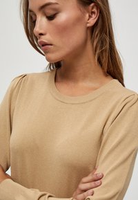 Beige neulepusero pyöreällä pääntiellä, neulotulla pinnalla ja poimutetuilla hihoilla, joka istuu napakasti ja on valmistettu pehmeästä kankaasta.