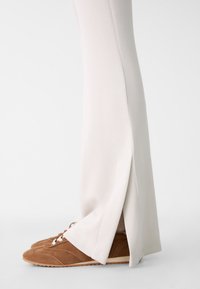 Světlé béžové sportovní kalhoty se bočními rozparky a hladkým povrchem, kombinované s hnědými semišovými botami s bílými tkaničkami a texturovanou podrážkou.