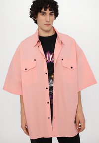 Camisa de manga corta de gran tamaño en rosa claro con botones negros y dos bolsillos en el pecho, llevada sobre una camiseta gráfica negra. Textura suave.