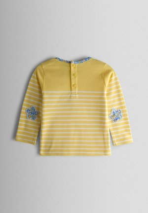 Dzeltens un balts svītrains krekls ar garām piedurknēm, zilām ziedu aplikācijām uz elkoņiem, pogu rāvējslēdzēju uz muguras un zilā ziedu apdares apkakli.