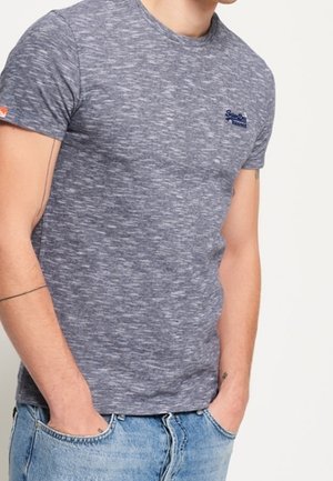 Mężczyzna w dopasowanym szarym melanżowym t-shircie z małym logo na piersi i jasnoniebieskich dżinsach, ręce w kieszeniach, na jednolitym tle.