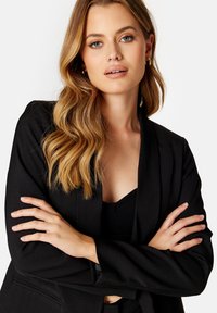 Zwarte blazer met een gestructureerde afwerking, voorzien van brede revers en een open voorkant. Model poseert met gekruiste armen en draagt een zwarte top eronder.