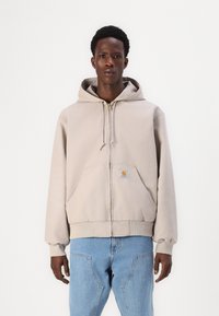Lichtbeige zip-up hoodie van zacht materiaal, met een voorste patch pocket en een capuchon met trekkoord. Draagt met lichtblauwe jeans.