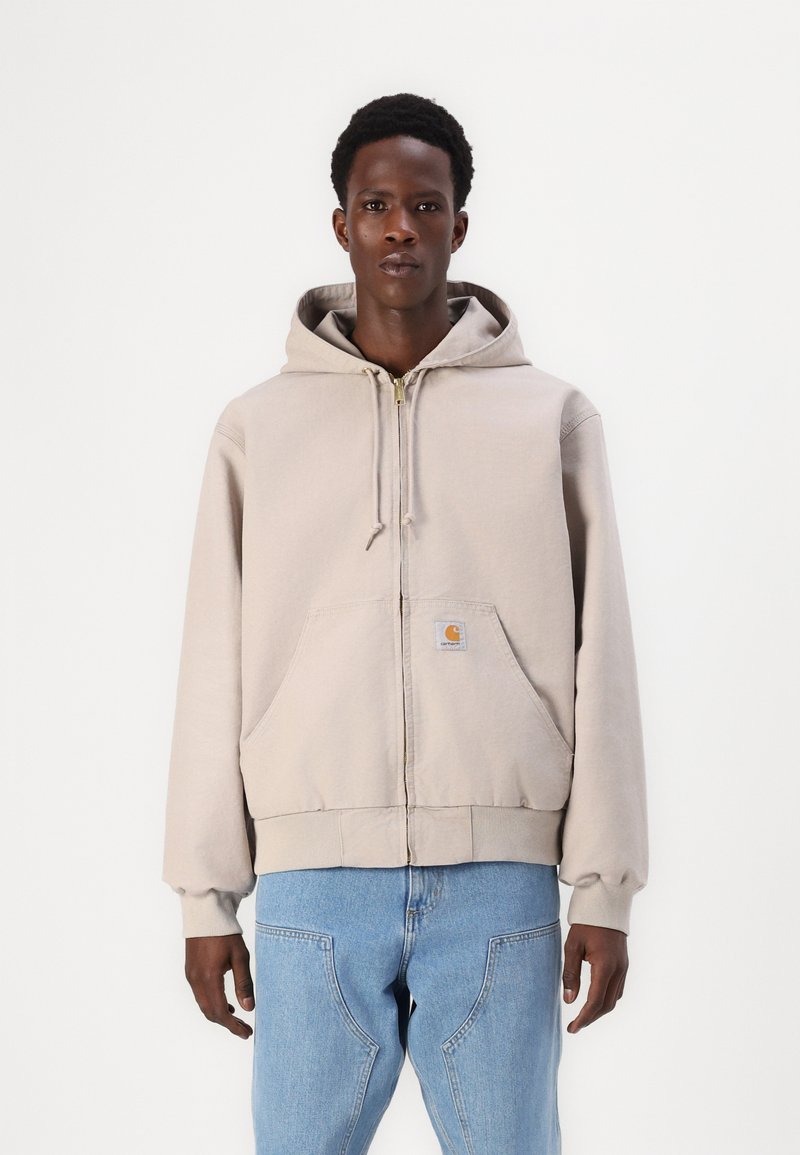 Lichtbeige zip-up hoodie van zacht materiaal, met een voorste patch pocket en een capuchon met trekkoord. Draagt met lichtblauwe jeans.