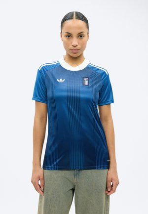 Moteris dėvinti mėlyną Adidas marškinėlį su baltu apykakle ir graikišku emblematu, stovinti atsukta į priekį prieš paprastą foną.