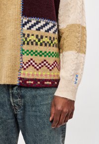 Kootud kampsun, millel on beeži, burgundia ja mitmevärviliste mustritega plaastrimuster. Iseloomulik palmikuteki struktuur ja sinised õmblusaktsendid.