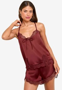 Frau trägt ein burgunderfarbenes Satin-Camisole und Shorts mit Spitzendetails, posiert mit einer Hand am Hals und der anderen am Oberschenkel.