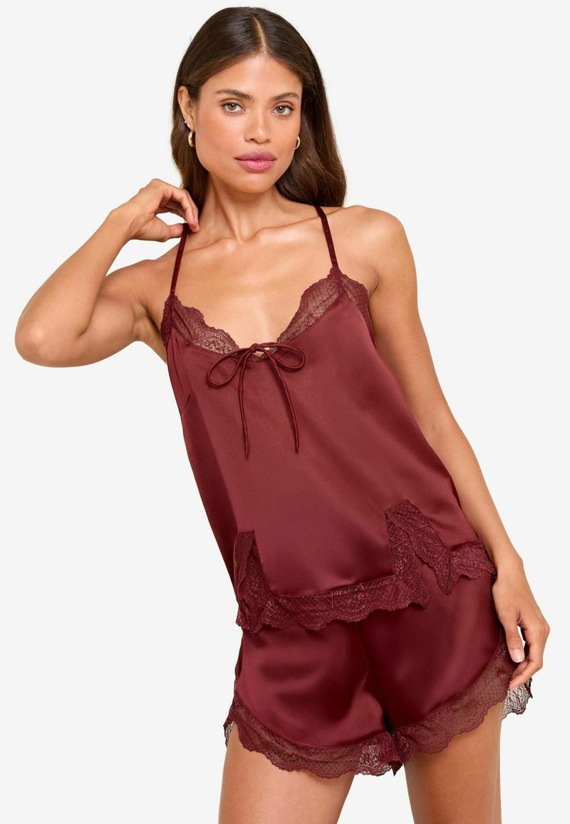 Frau trägt ein burgunderfarbenes Satin-Camisole und Shorts mit Spitzendetails, posiert mit einer Hand am Hals und der anderen am Oberschenkel.