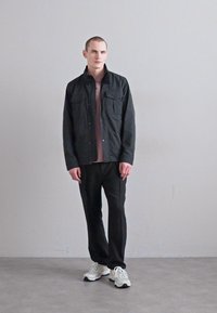 Jachetă neagră cu nasturi, cu două buzunare pe piept, purtată peste o cămașă de culoare mov deschis. Pantaloni negri și adidași albi completează ținuta.