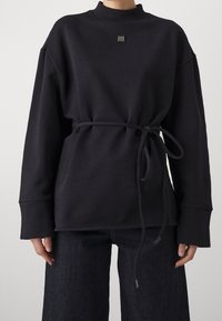 Felpa nera a maniche lunghe con collo rotondo, cintura legata in vita e un piccolo logo quadrato sul petto, indossata con jeans scuri.