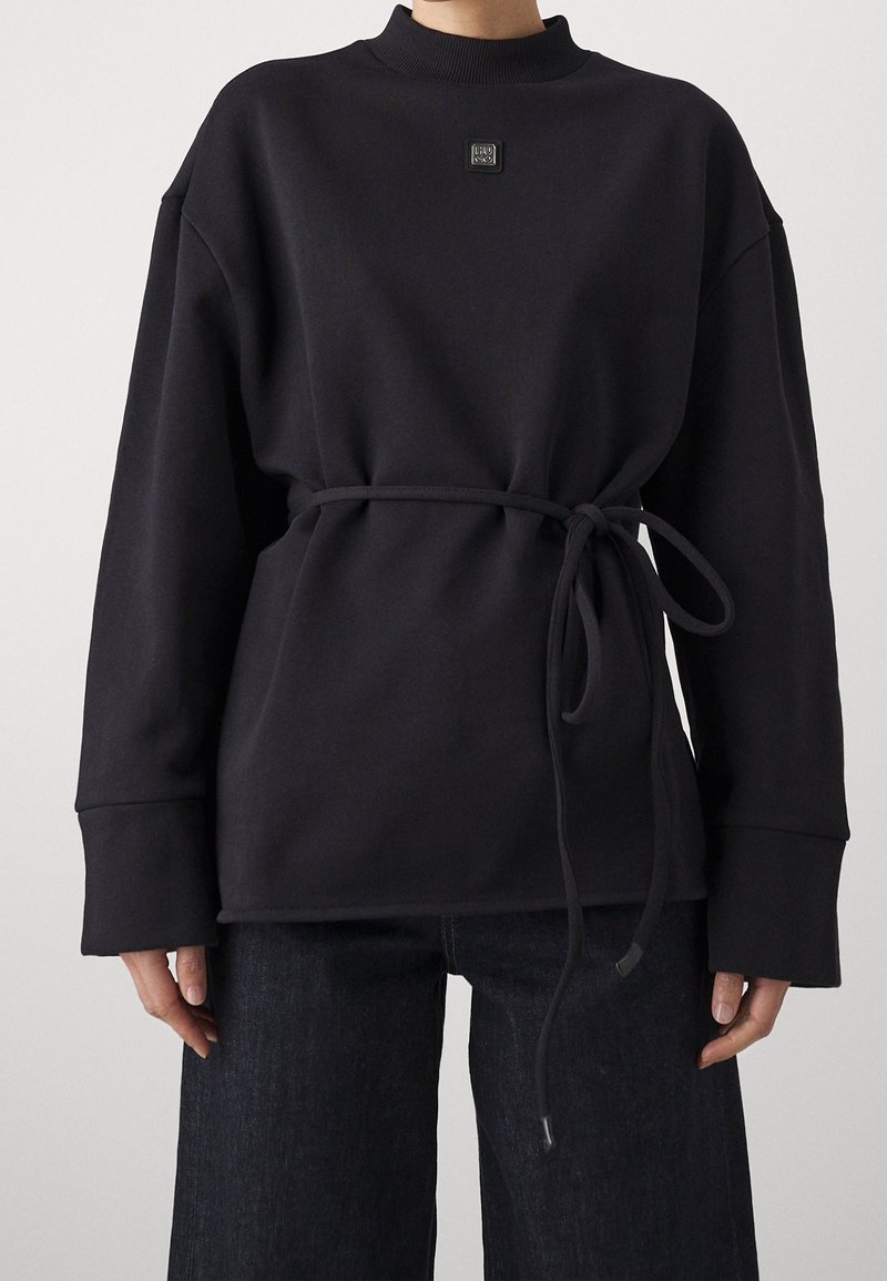 Felpa nera a maniche lunghe con collo rotondo, cintura legata in vita e un piccolo logo quadrato sul petto, indossata con jeans scuri.