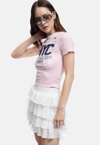 Rosa kortärmad topp med texturerad yta och NYC-tryck, parat med en vit lager på lager volangkjol. Modellen bär solglasögon.