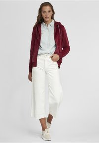 Burgunderfarget zip-up hettejakke over en lys blå stripete knappskjorte, kombinert med vide hvite capribukser og beige sneakers.