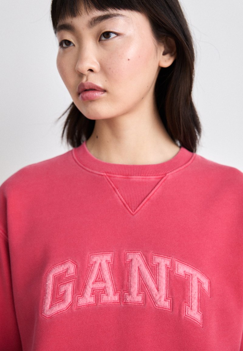 Różowy sweter z logo "GANT" w teksturze i odważnej czcionce. Posiada okrągły dekolt oraz miękką tkaninę w luźnym kroju.
