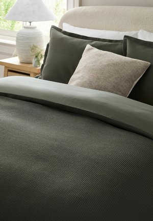Seng med olivengrønt tekstureret sengetøj, beige pude, cremefarvet sengegavl, træ natbord, hvid lampe og en lille plante nær vinduet.