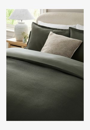 Bett mit olivgrüner, strukturierter Bettwäsche, beigem Kissen, cremefarbenem Kopfteil, hölzernem Nachttisch, weißer Lampe und kleiner Pflanze neben dem Fenster.
