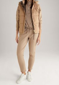 Chaleco acolchado beige con patrón acolchado, usado sobre un conjunto beige a juego. Cuenta con un cuello con cremallera y cintura con cordón, combinado con zapatillas blancas.