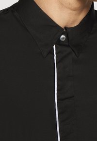 Camicia nera con colletto button-down, caratterizzata da una striscia verticale bianca lungo la pattina e un singolo bottone argentato al colletto.