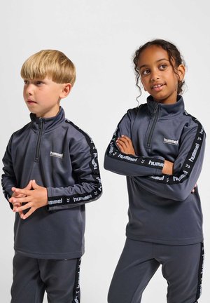 To børn iført matchende mørkegrå hummel træningsdragter med mærkede striber, står side om side mod en lys baggrund.