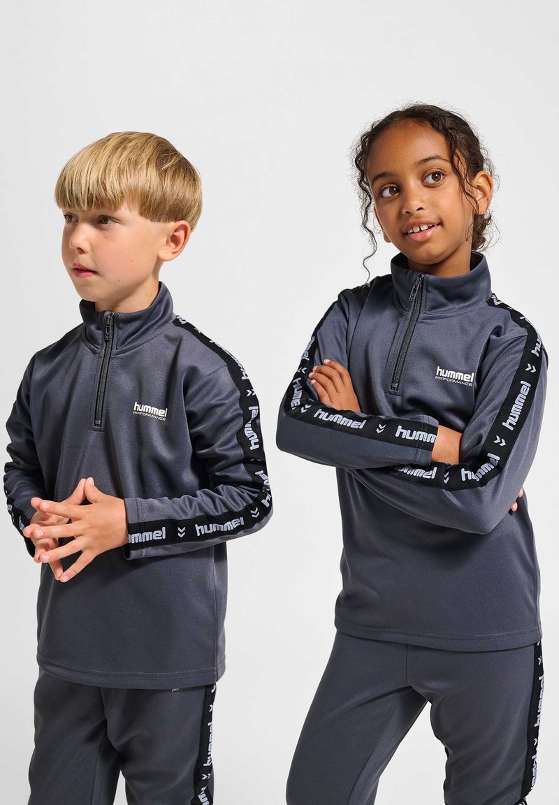 To børn iført matchende mørkegrå hummel træningsdragter med mærkede striber, står side om side mod en lys baggrund.