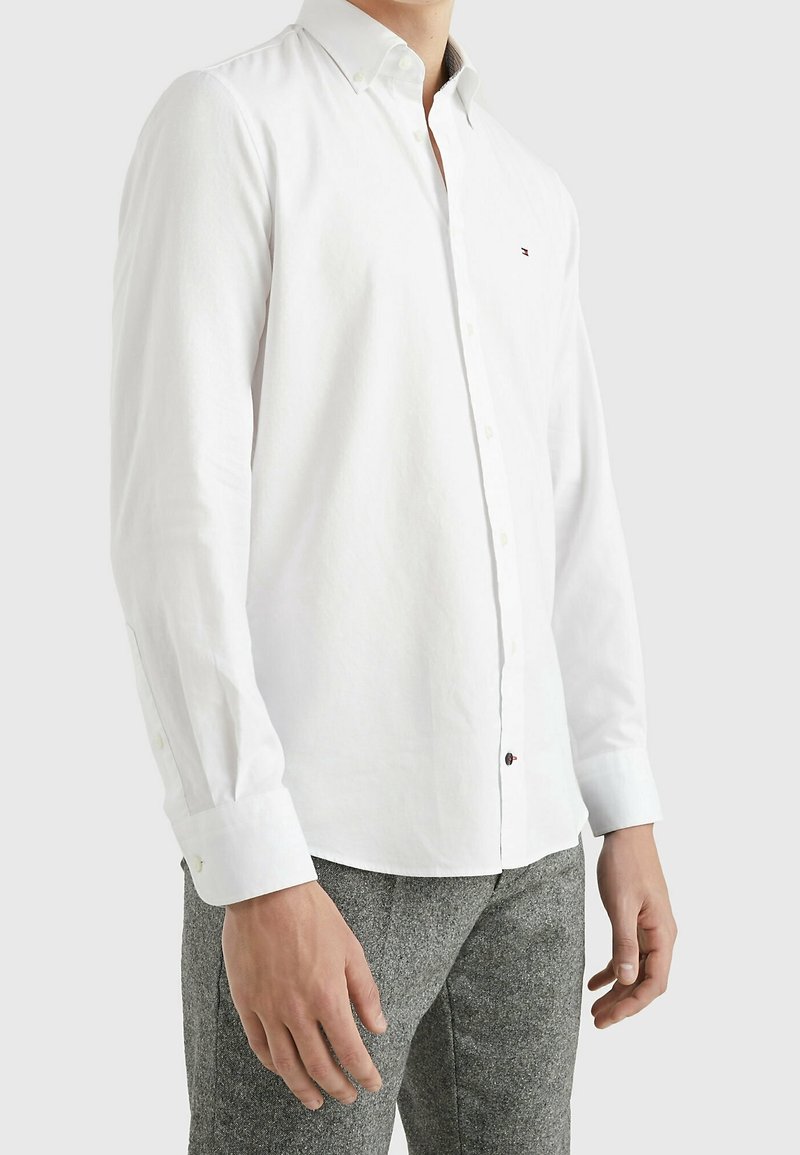 Wit overhemd met knopen, een gestructureerde kraag, lange mouwen en een subtiele textuur; heeft een klein logo bij de borst en bruine knopen.