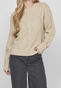 Beige kabelstickad tröja med diamant- och ribbmönster, rund halsringning och avslappnad passform; ihop med mörkgrå jeans.
