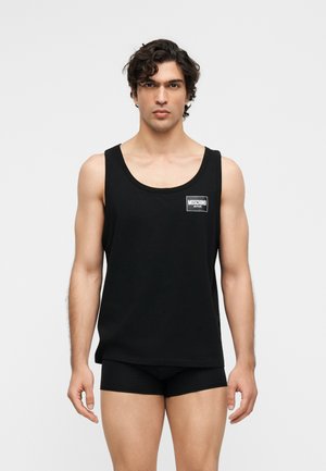 Άνδρας με κοντά σγουρά μαλλιά, φορώντας μαύρο αμάνικο top Moschino Active και μαύρα εσώρουχα, στέκεται μπροστά σε έναν απλό φωτεινό φόντο.