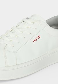 Λευκό sneaker χαμηλού πάτου με λευκά κορδόνια και κόκκινη σήμανση "HUGO" στο πλάι, με καθαρό, μινιμαλιστικό σχεδιασμό.