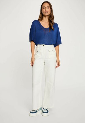 Donna con top blu a maniche corte, pantaloni bianchi a vita alta e gamba larga con tasche, e sneakers platform blu e bianche, in piedi.