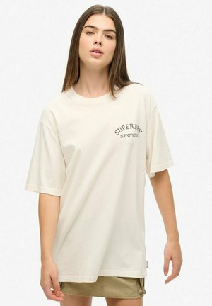Kremowa bawełniana koszulka z krótkim rękawem, z okrągłym nadrukiem "SUPERDRY NEW YORK" w ciemnym kolorze na lewej części klatki piersiowej.