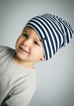Junges Kind lächelt, trägt eine marineblaue und weiß gestreifte Mütze und ein hellbeiges Langarmshirt vor einem neutral grauen Hintergrund.