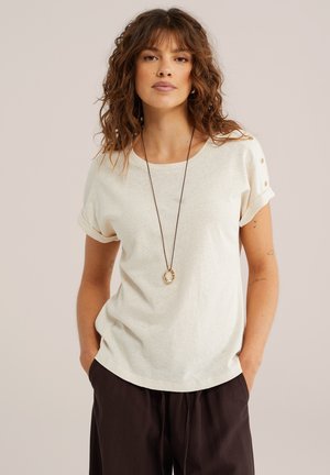 Mujer con cabello castaño ondulado, vestida con una camisa corta de manga corta color crema con detalles de botones, pantalones oscuros y un collar largo con colgantes circulares.